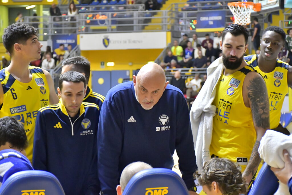 Coach Boniciolli della Reale Mutua Basket Torino dà indicazioni alla squadra durante un timeout, circondato dai giocatori in divisa gialla, sullo sfondo del Pala Gianni Asti.